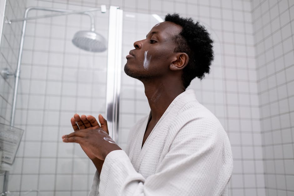 African American man in bathrobe applying skincare cream in a bathroom.