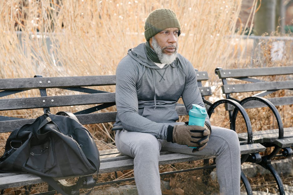 Senior man in gray hoodie sitting on a bench holding a tumbler outdoors in fall.