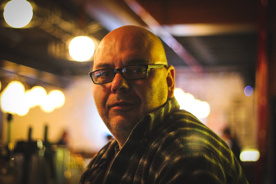 Bald man wearing glasses in a warm-lit café, captured with shallow depth of field.
