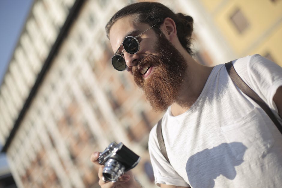 Happy bearded man with sunglasses holding a camera and smiling outdoors in a city.