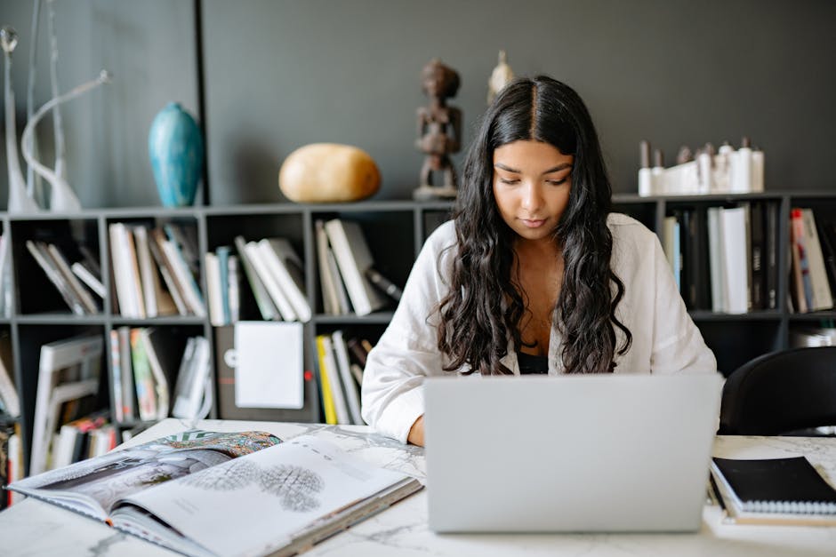 A woman with long hair works on her laptop at a desk with books and a bookcase behind her.