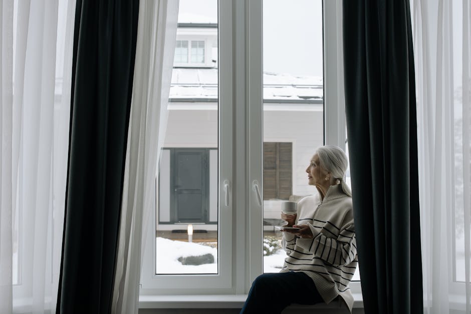 Elderly woman sitting by window, enjoying a warm drink in a cozy winter setting.