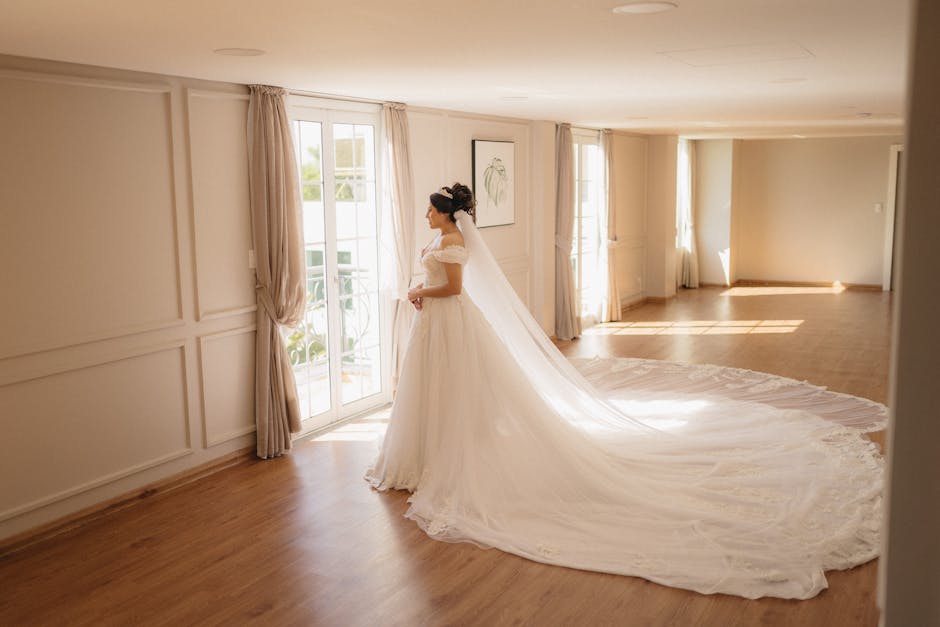 A beautiful bride stands gracefully by a window in a luxurious indoor setting.