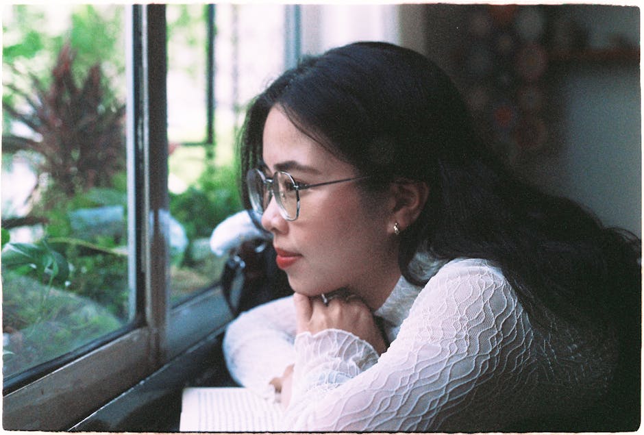 Young woman in glasses and a white blouse looks thoughtfully outside through a window.