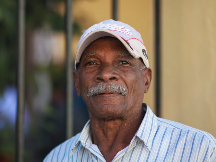 Headshot of a senior man wearing a cap and striped shirt outdoors. Calm expression.